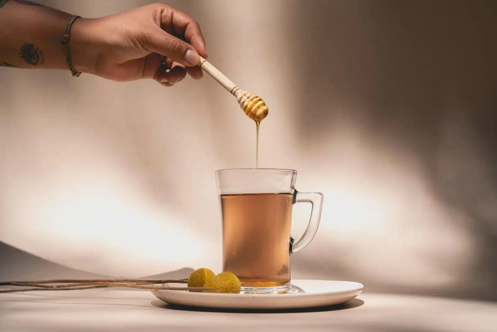 一隻手用沾蜜棒將蜂蜜滴入玻璃杯熱茶中，溫水沖泡是蜂蜜常見的吃法