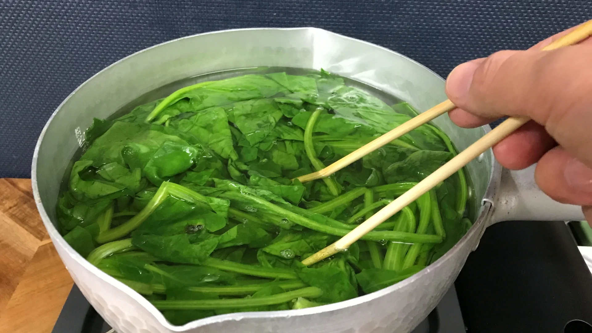 瓦斯爐上的鍋子裡正在汆燙菠菜，一雙筷子把菜壓進滾水中