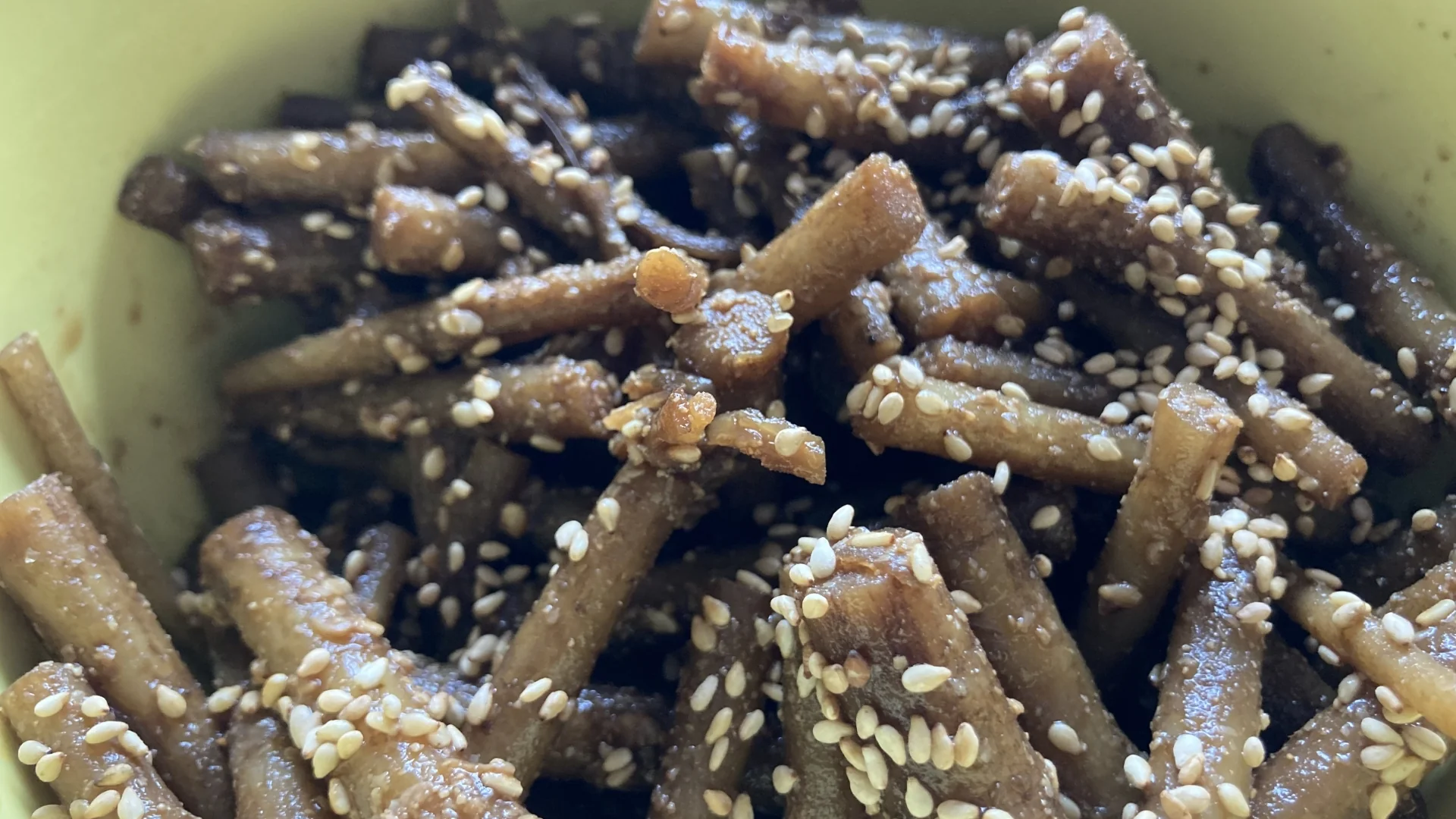 醬燒牛蒡條收汁後撒上大量白芝麻，盛在淺綠色碗裡，表面帶著醬色的光澤