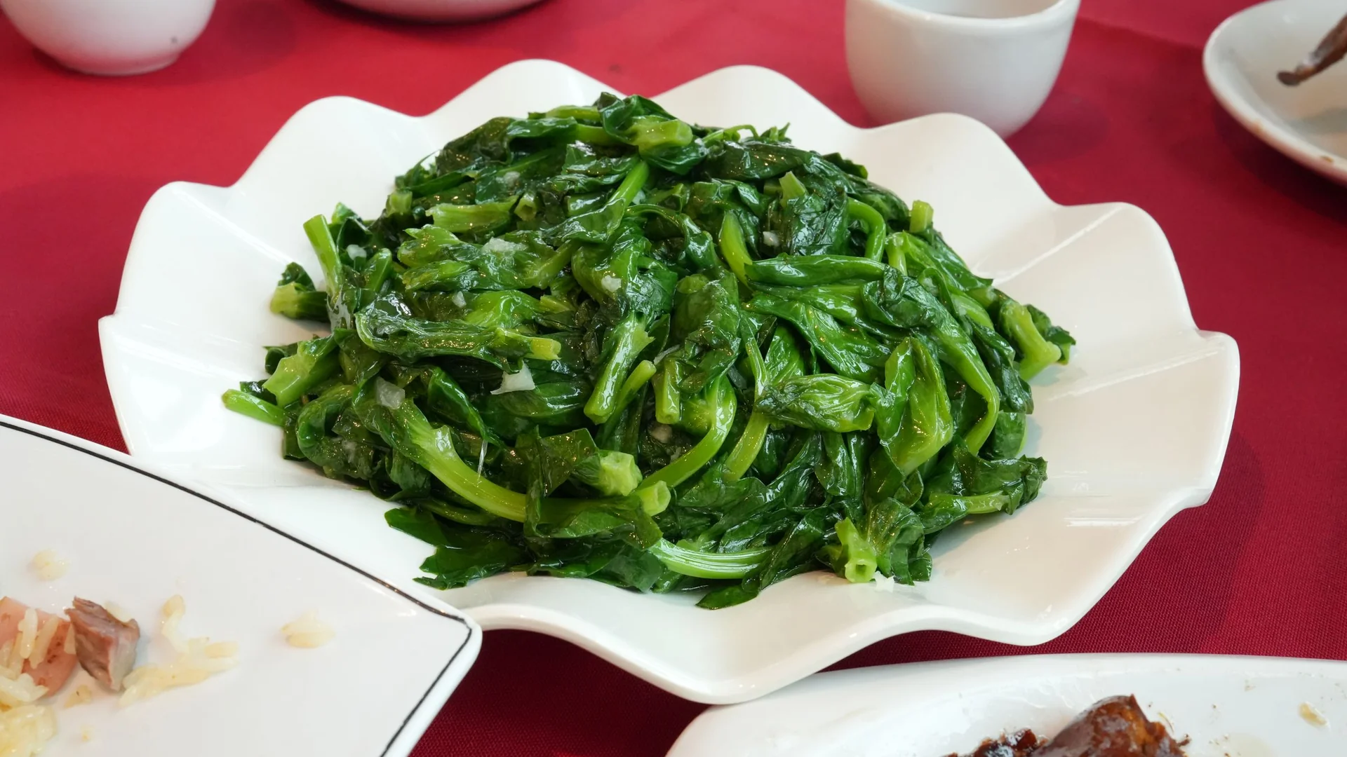 白色花邊盤上炒得油亮的綠色葉菜，放在紅色桌布的中式餐桌上